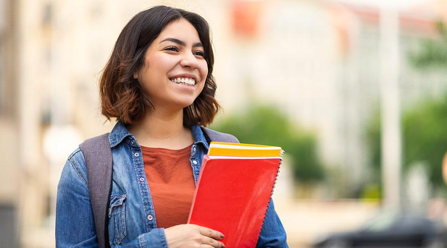年轻的拉丁裔女学生背着背包，手里拿着笔记本，面带微笑